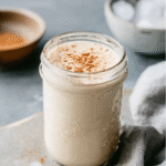 Glass mason jar filled with a creamy vanilla protein shake, topped with cinnamon, styled on a gray stone counter with ice cubes, linen cloth, and a bowl of cinnamon in the background.