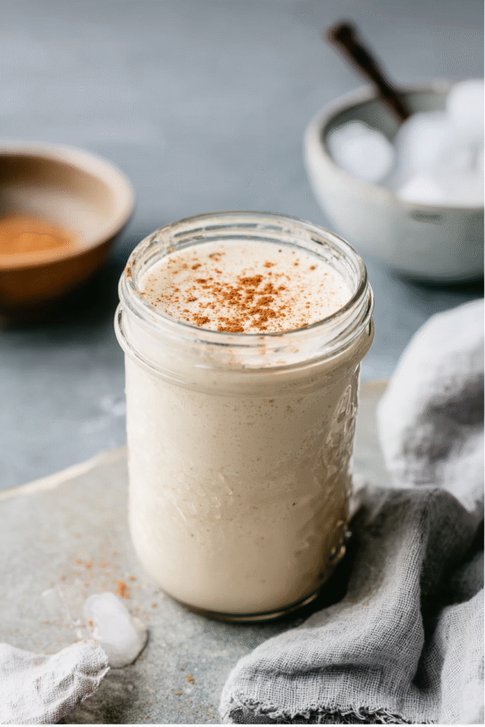 Vanilla Protein Shake That Actually Tastes Good – 2025 1 Glass mason jar filled with a creamy vanilla protein shake, topped with cinnamon, styled on a gray stone counter with ice cubes, linen cloth, and a bowl of cinnamon in the background.