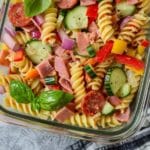 Overhead shot of high protein Italian pasta salad in a glass container with rotini pasta, sliced cucumber, red onion, turkey pepperoni, bell peppers, and fresh basil.