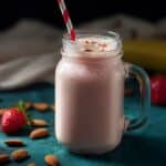 Low calorie protein shake in a mason jar with a striped straw, surrounded by almonds, strawberries, and banana on a teal surface.